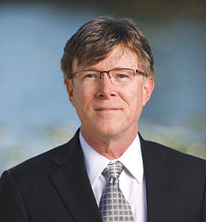 Man in dark suit, white shirt and gray tie wearing glasses and smiling