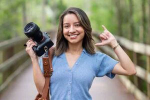 A smiling woman with shoulder-length brown hair in a blue, short-sleeved button-up sweater holds a camera in her right hand and gives a Wings Up! gesture with her left hand