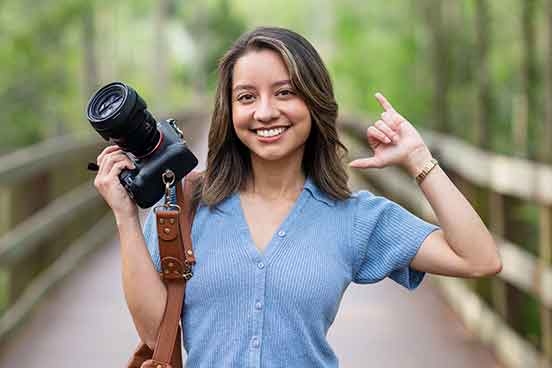 A smiling woman with shoulder-length brown hair in a blue, short-sleeved button-up sweater holds a camera in her right hand and gives a Wings Up! gesture with her left hand
