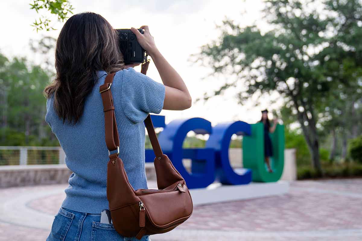 A photographer, as viewed from behind, takes photos of a woman in a graduation cap standing in the blue and green FGCU logo on the Library Lawn