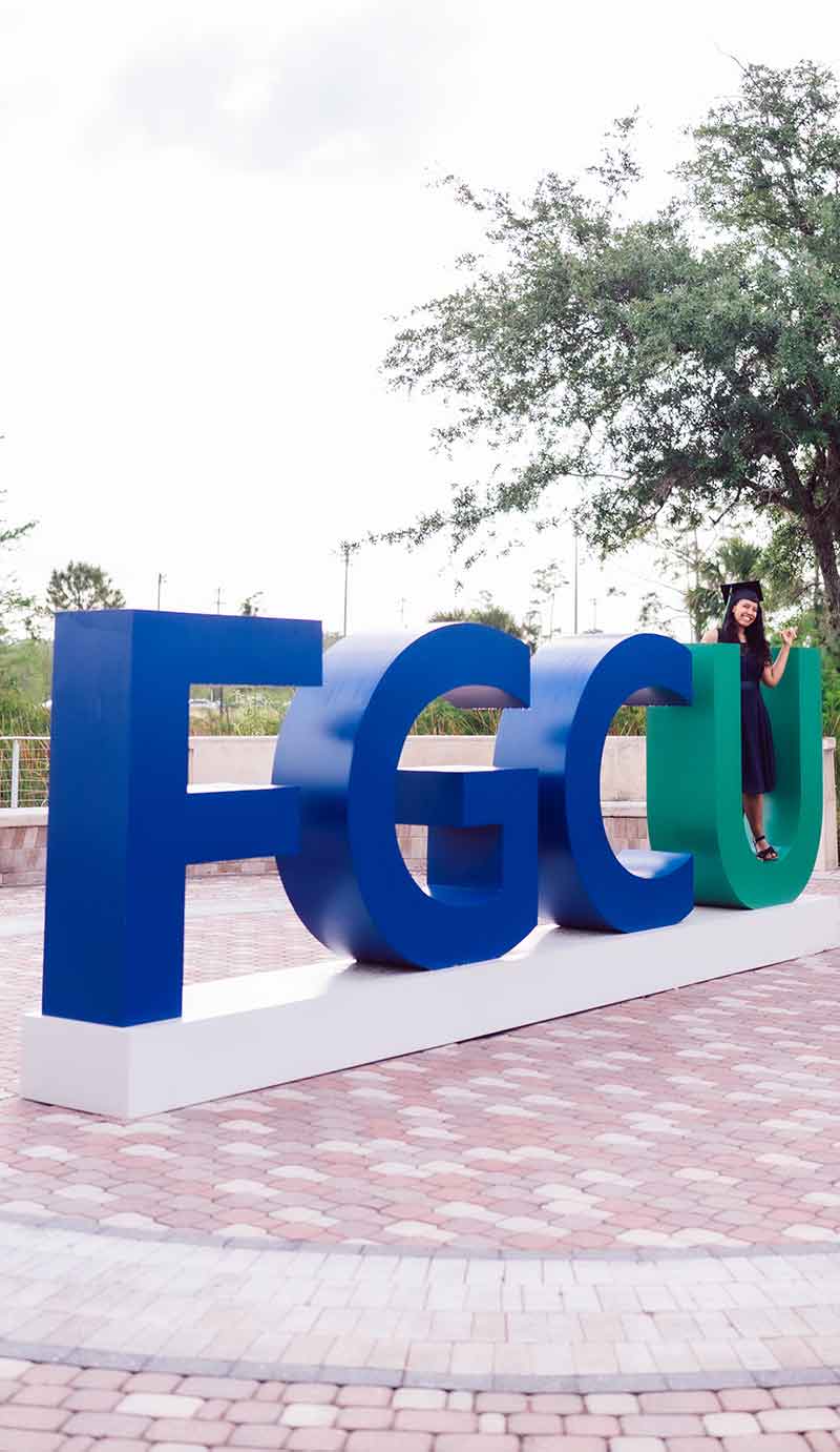 Large blue and green FGCU logo on Library Lawn with a woman standing inside the letter U