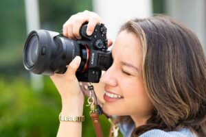 A brown-haired woman in a blue sweater holds a camera up to her right eye to take a photo while her left eye is closed