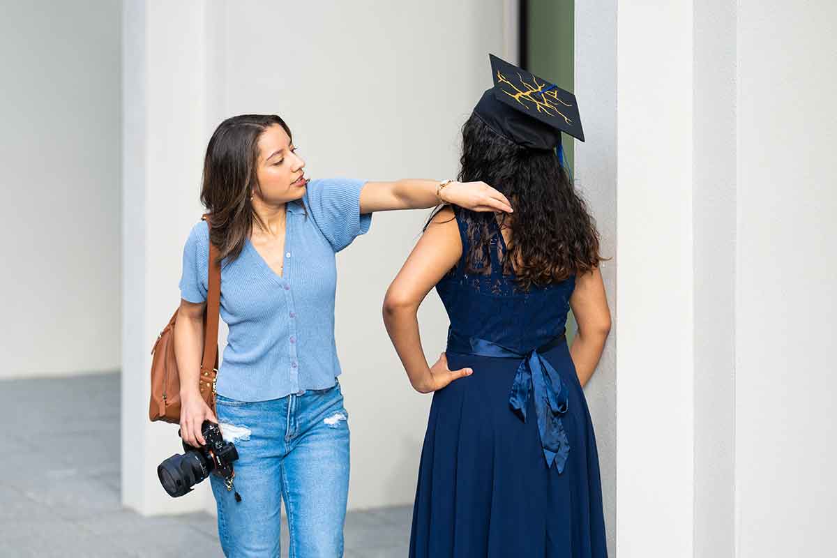 A photographer in a blue sweater fixes the hair of a woman in a navy blue dress and mortarboard graduation cap standing with her back to the camera