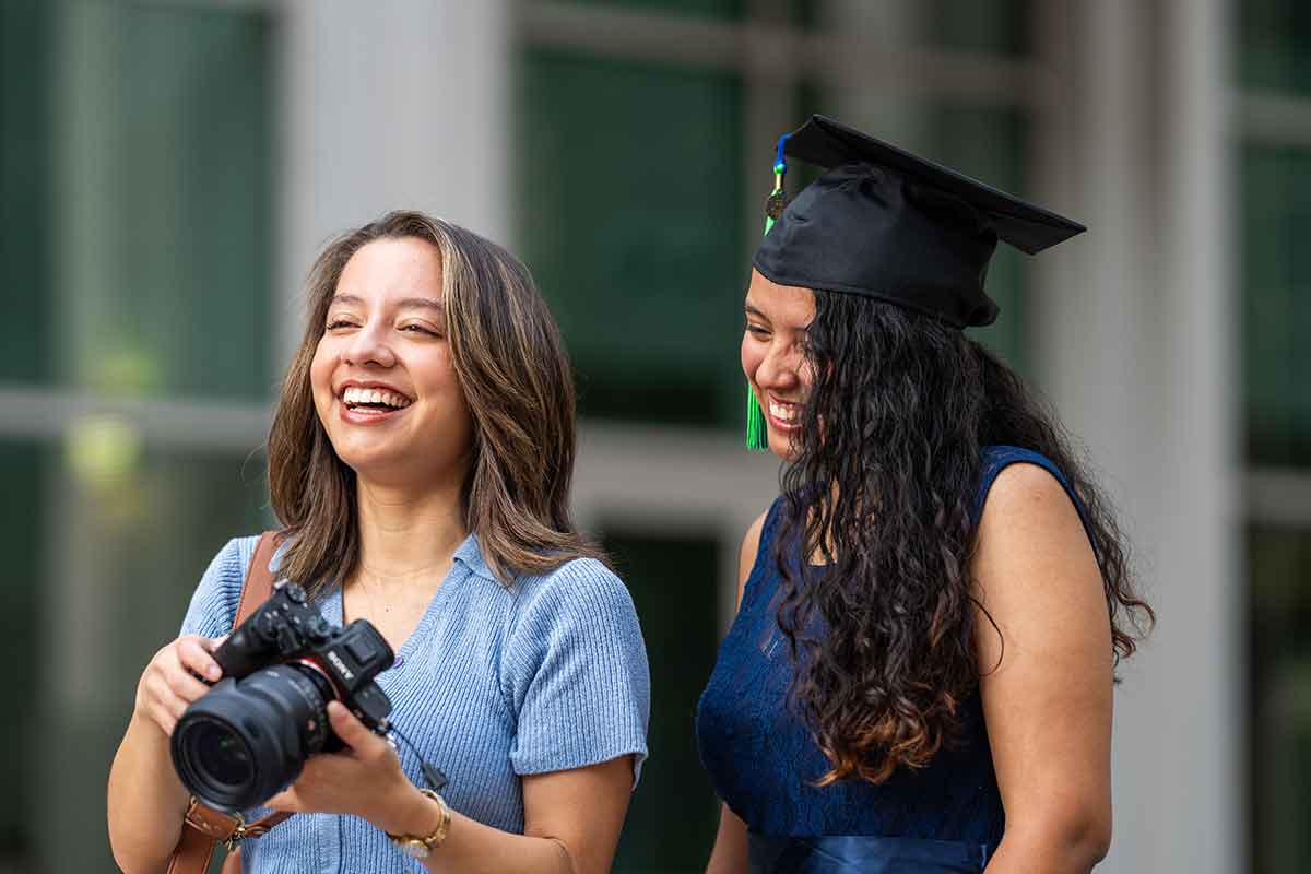 A laughing woman in a blue sweater shows a woman in a navy blue dress and a mortarboard graduation cap a photo on her camera