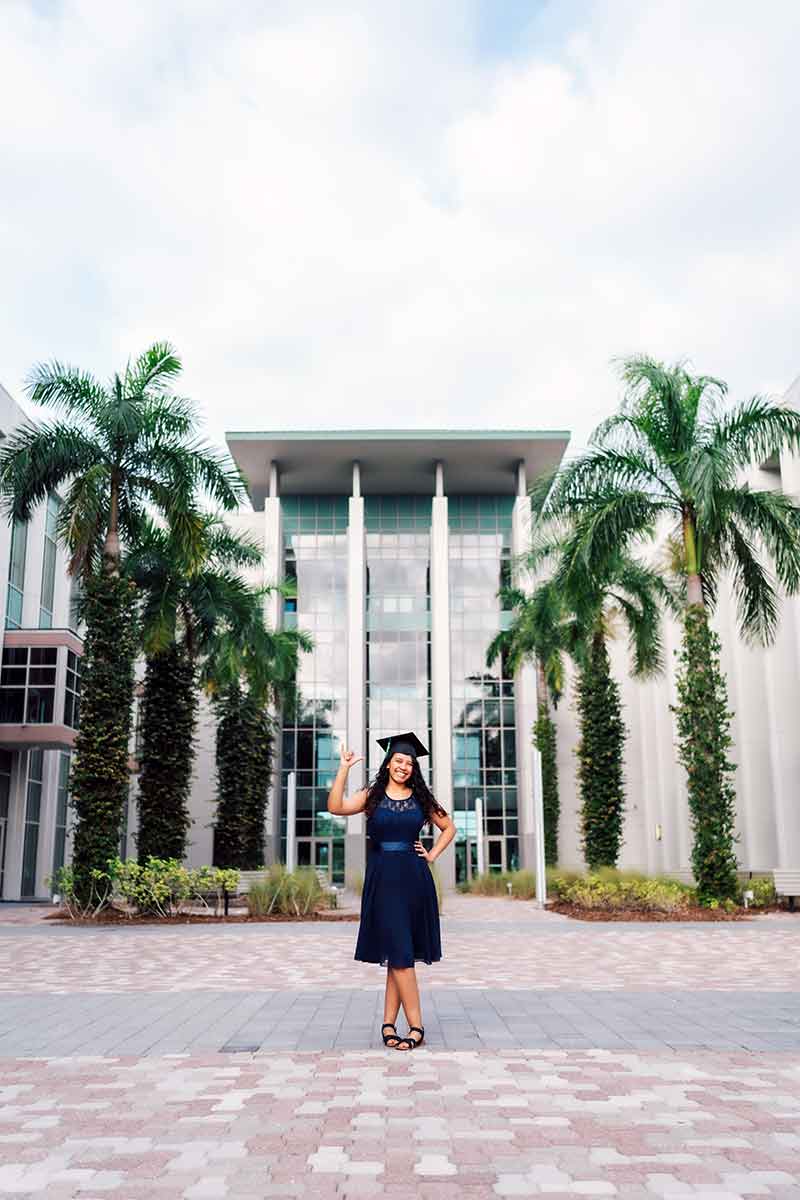 A woman in a navy blue dress stands facing the photographer with her feet crossed while wearing a mortarboard and making a Wings Up! gesture with her right hand