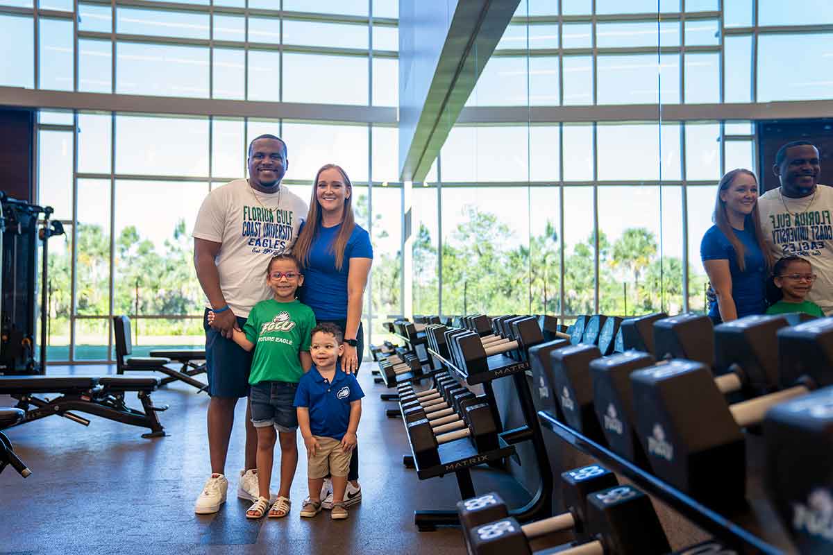 A family of four wears FGCU colors in a fitness center with workout equipment along the mirrored right wall
