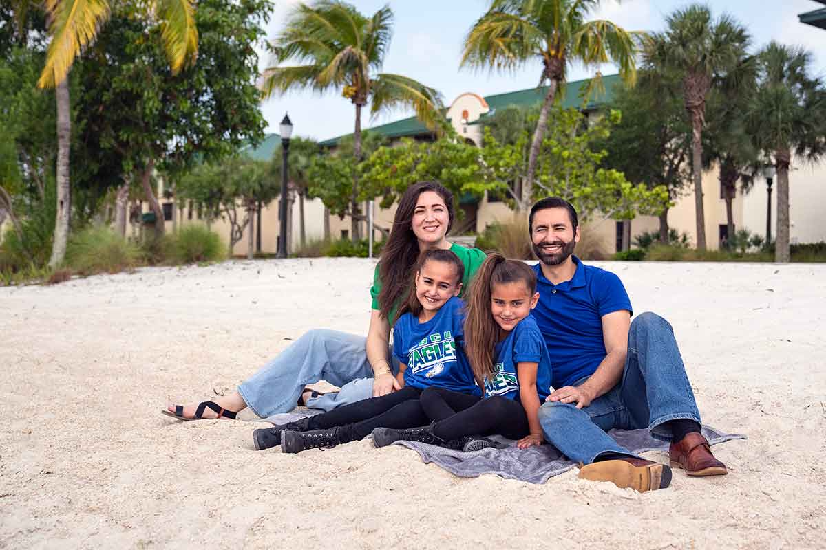 Two adults and two children dressed in FGCU green and blue sit on a sandy beach in front of residential housing and palm trees