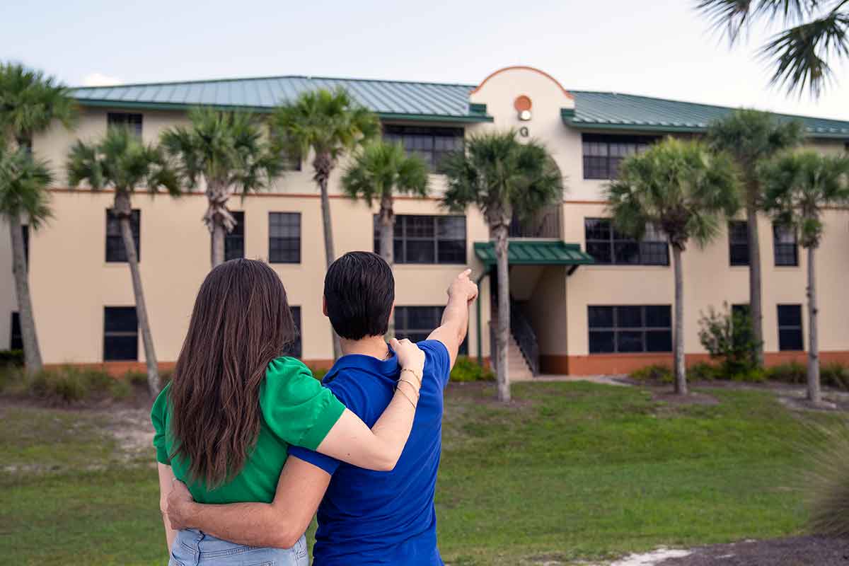 A woman in a green top and jeans stands with a man in a blue top and jeans, facing away from the camera. He's pointing to a green-roofed building lined with palm trees.