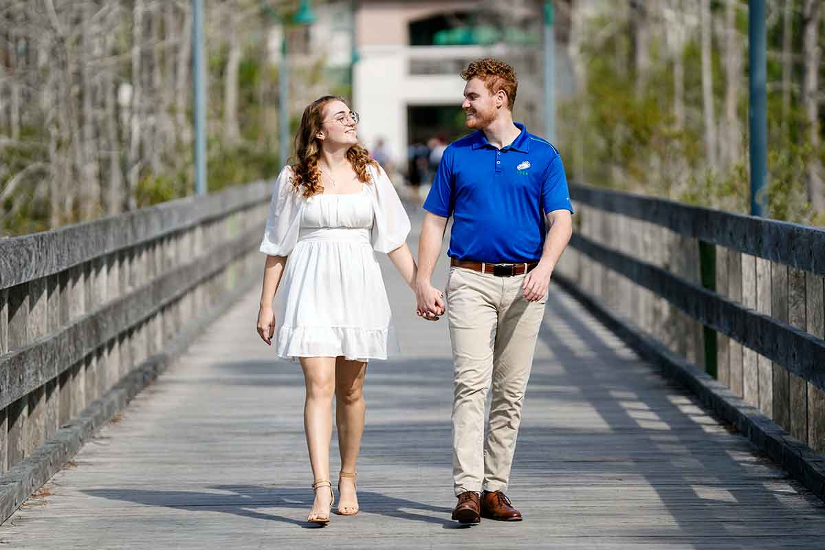 A woman in a short white dress and a man in a blue polo shirt hold hands and look at each other as they walk along a wooden boardwalk