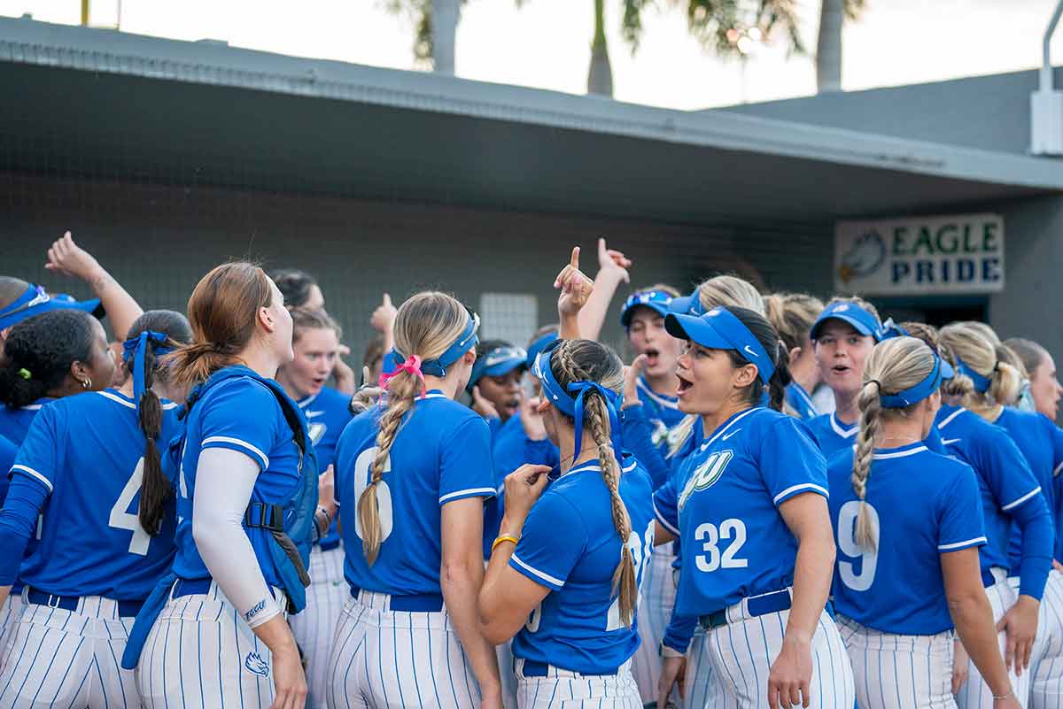 A group of softball players in blue jerseys and white and blue striped pants celebrate in front of a dugout