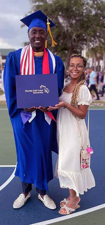 A man in blue cap and gown stands next to a woman in a white dress, holding his diploma together
