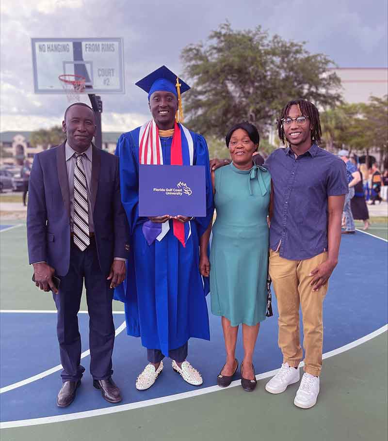 A student in blue graduation cap and gown, holding his diploma. He stands on a basketball court with three family members.