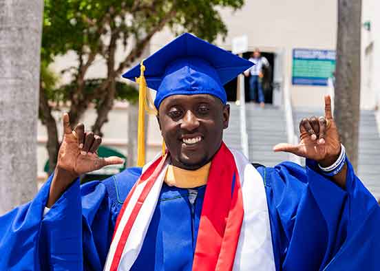 A smiling student in blue graduation cap and gown with both hands in a Wings Up gesture