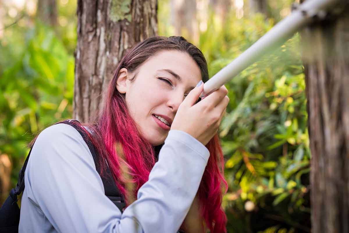 A woman with red-tipped brown hair looks into a white PVC pipe attached to a tree.