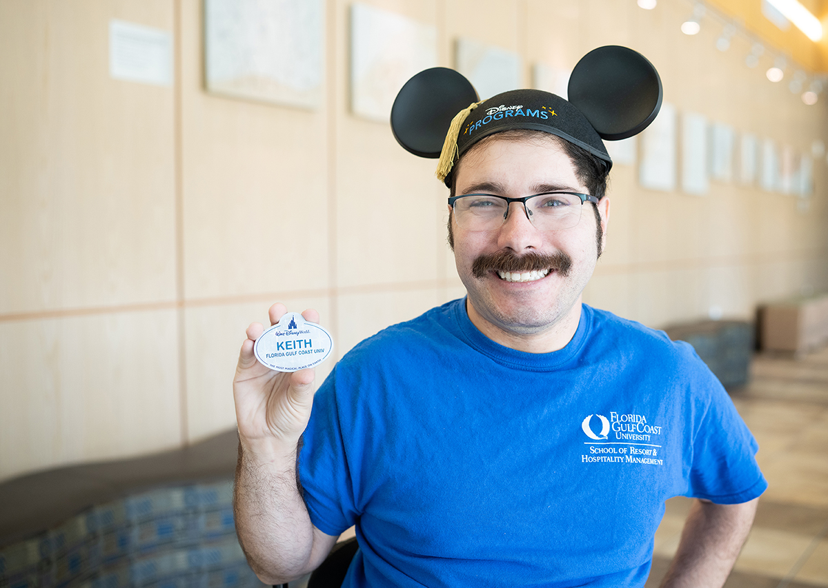 A man with Disney ears, glasses and a moustache in a blue, short-sleeved shirt sits in a wheelchair, smiling and holding up a Disney nametag with the name Keith and the hometown Florida Gulf Coast University