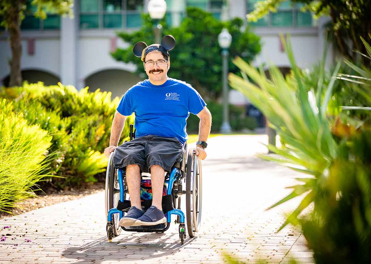 A man with Disney ears, glasses and a moustache in a blue, short-sleeved shirt sits in a wheelchair outdoors
