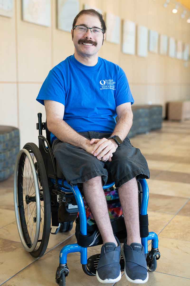 A man with glasses and a moustache in a blue, short-sleeved shirt sits in a wheelchair