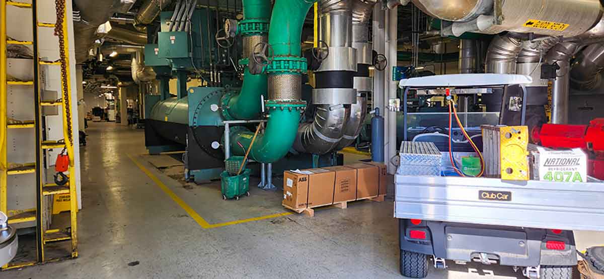 Large green and silver condensers inside of a building, next to a golf cart filled with maintenance equipment, including a ladder