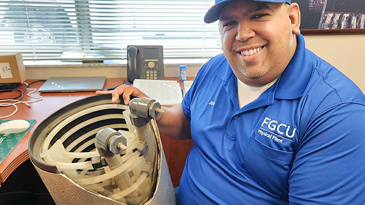 A smiling man in a blue polo shirt and hat holds a cylinder with exposed inner workings