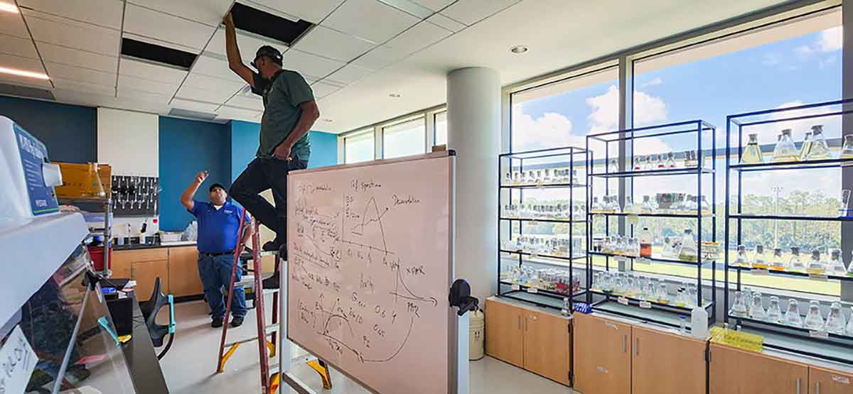 One man on a ladder and one below work in a classroom on HVAC