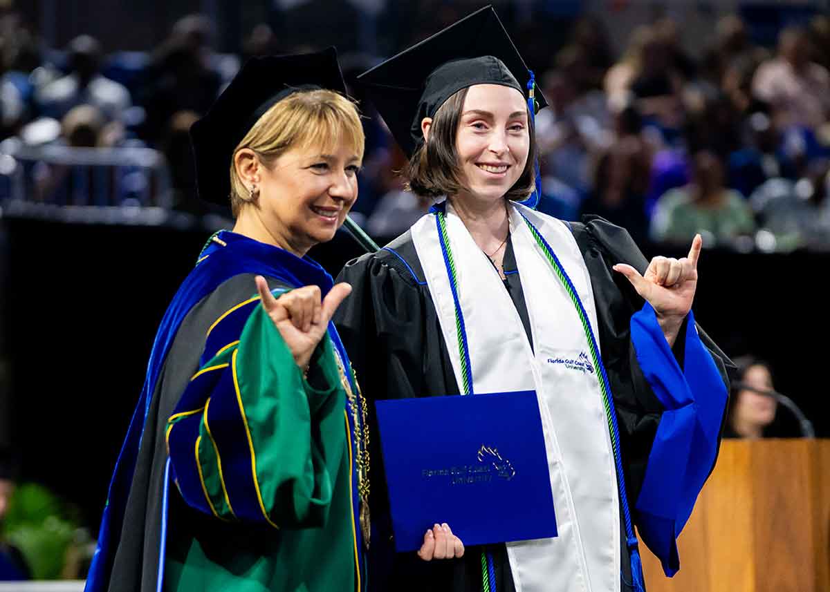 A blonde woman in cap and gown and a brunette woman receiving her FGCU diploma give the Wings Up gesture.