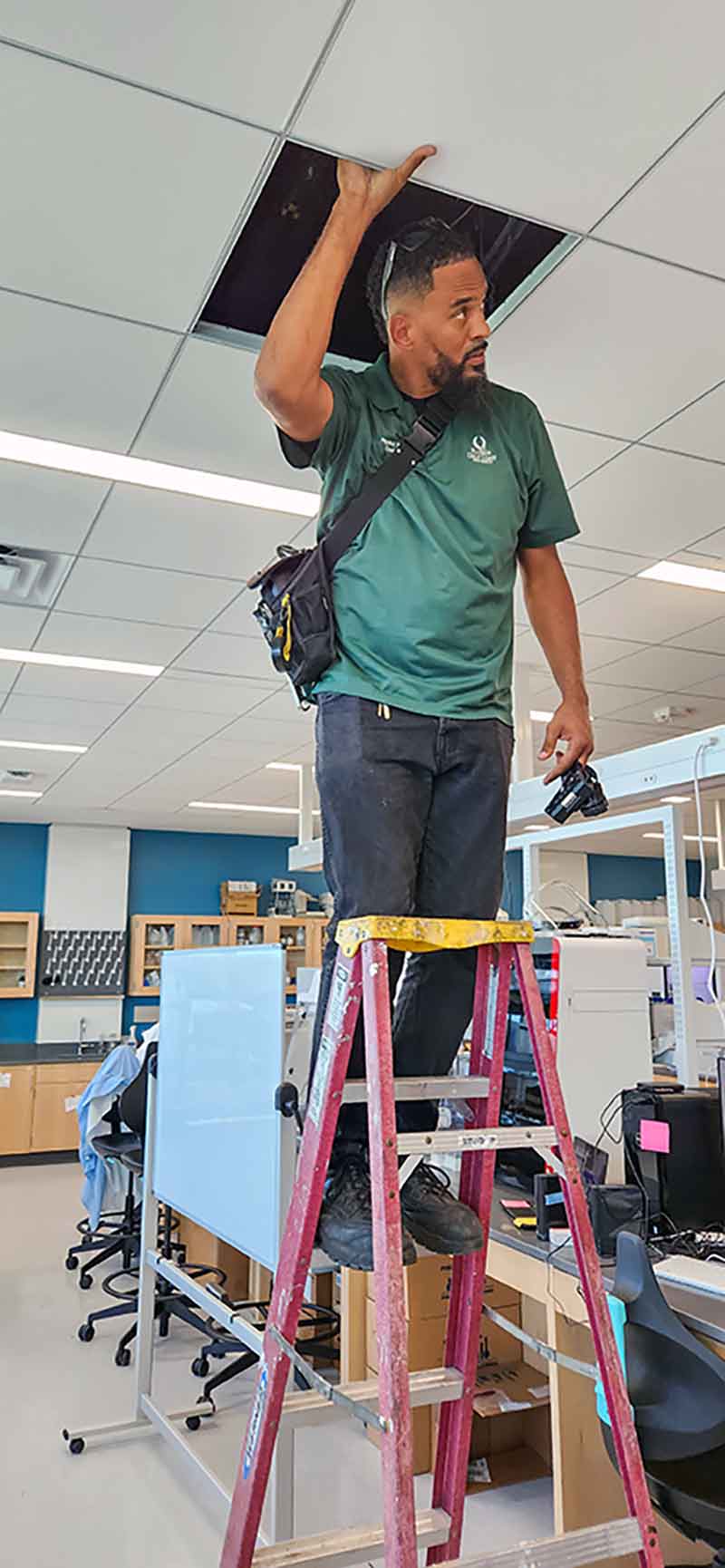 A man in a green shirt on a ladder looks inside the ceiling of a classroom