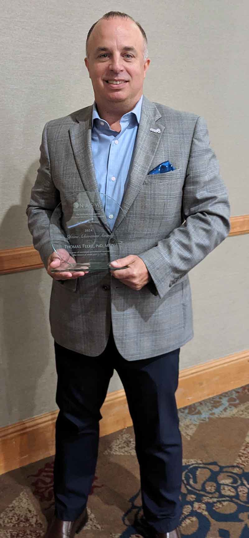 A smiling man in a blue button-up shirt, grey suit jacket and black pants holds a glass award.