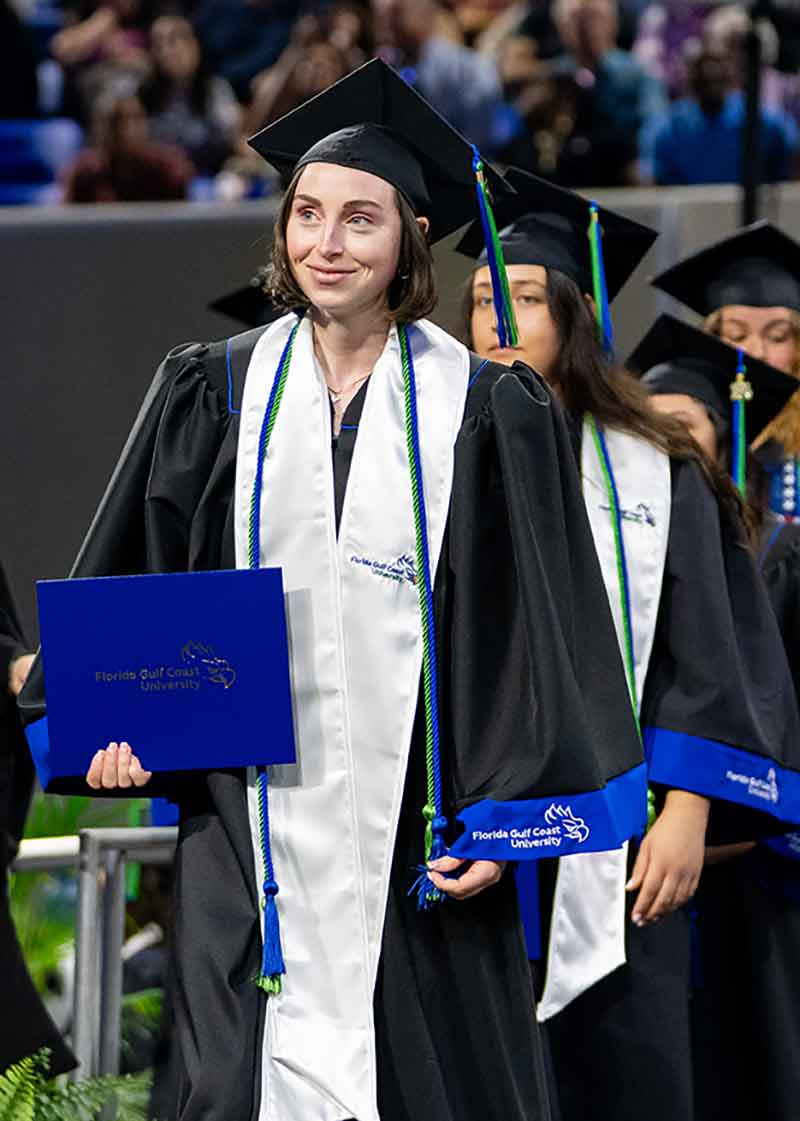 A smiling woman in black cap and gown with a green tassel, white stole and blue and green honor cords walks on a graduation stage, her diploma in her right hand.