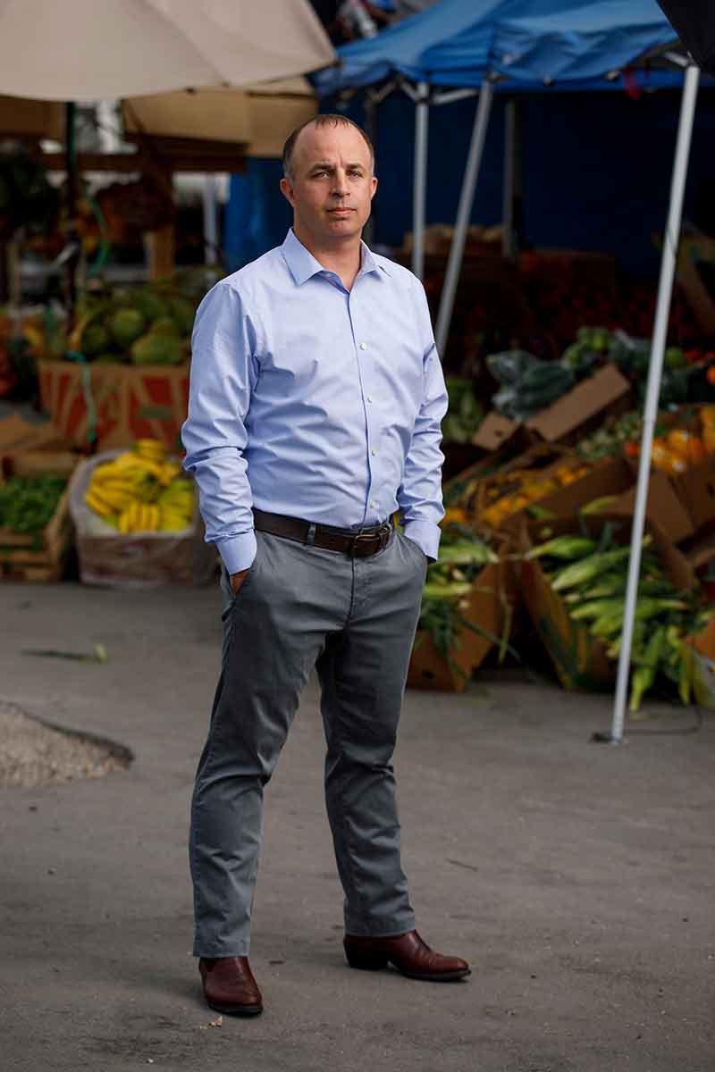 A man in a light blue button-up shirt and grey slacks stands in front of a market.