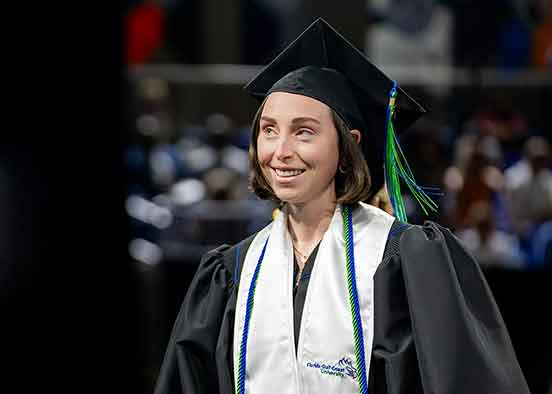 A smiling woman in black cap and gown with a green tassel, white stole and blue and green honor cords.