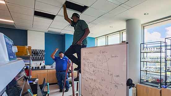 One man on a ladder and one below work in a classroom on HVAC