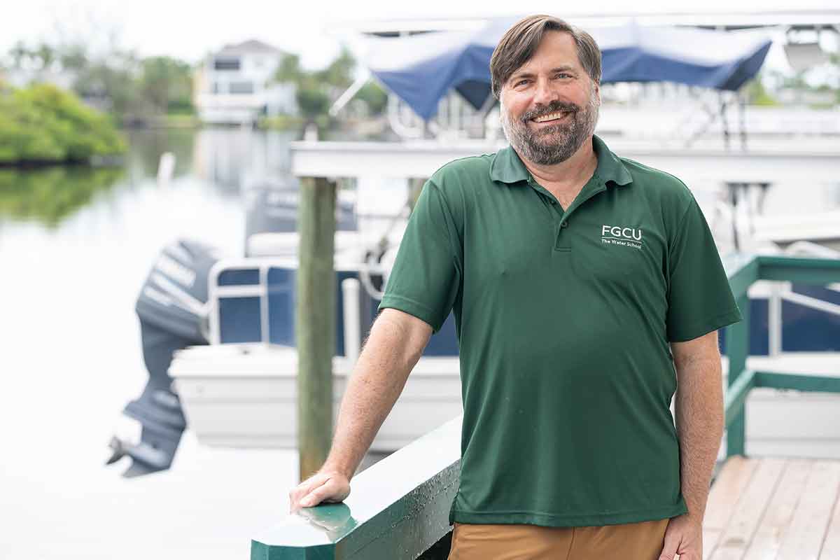 A smiling man in a green Polo shirt stands with one hand on a railing, a boat visible behind him