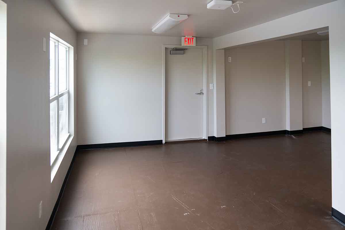 An empty room with white walls, a window, a side exit door and plain, brown flooring.