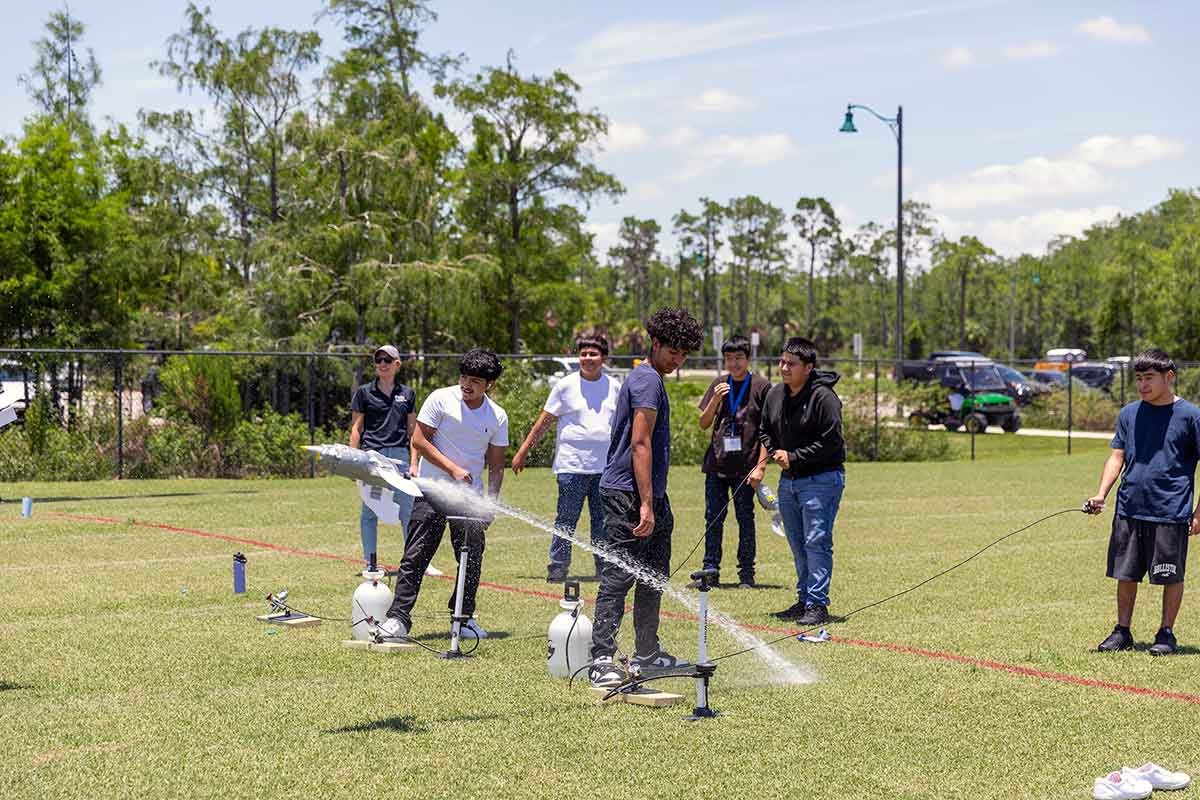 A group of young men outdoors watch a homemade rocket, featured in mid-flight