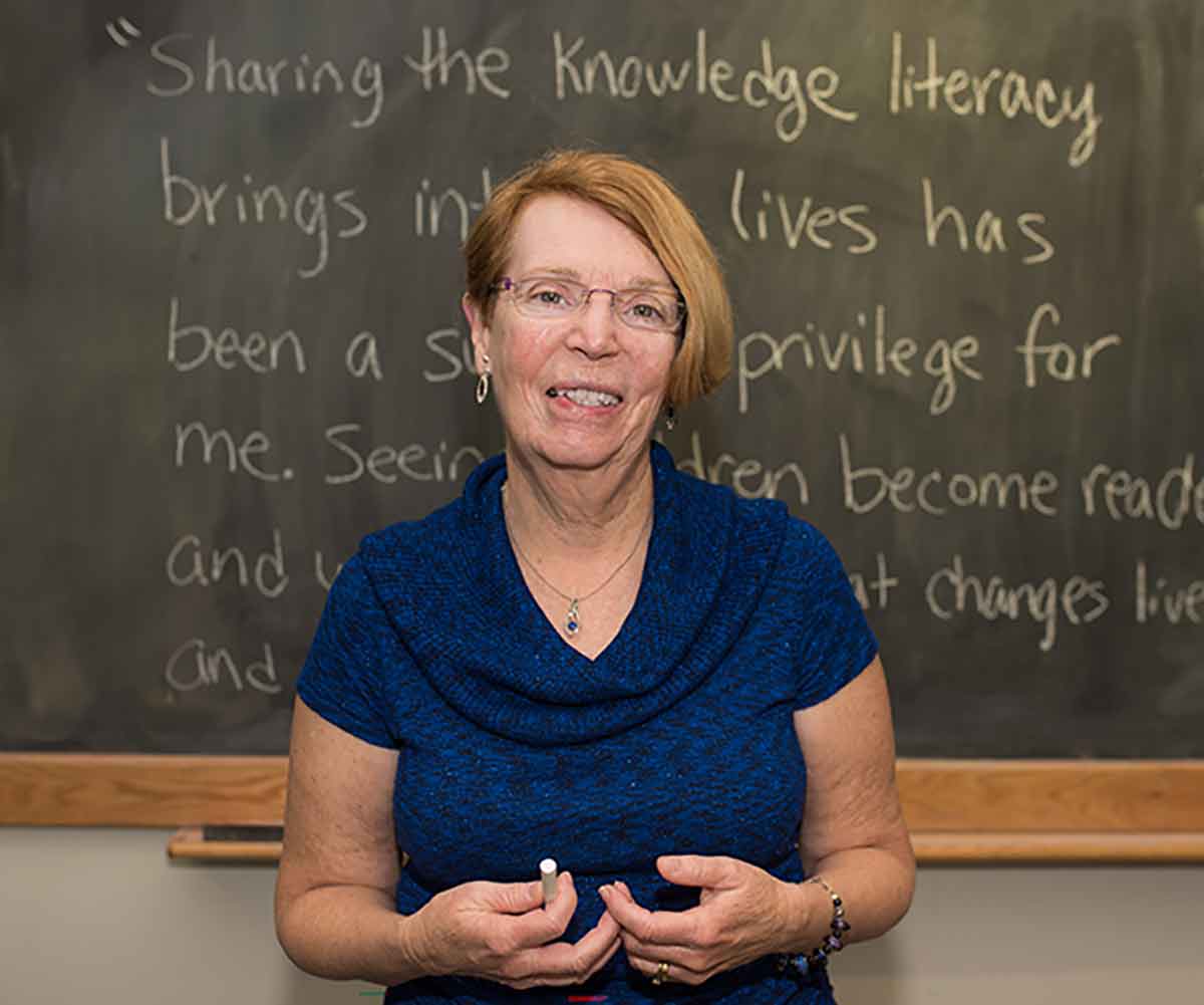 A woman in a blue short-sleeved sweater stands in front of a chalkboard. Part of a quote is visible behind her: "Sharing the knowledge literacy brings into lives has been a singular privilege for me. Seeing children become readers..."