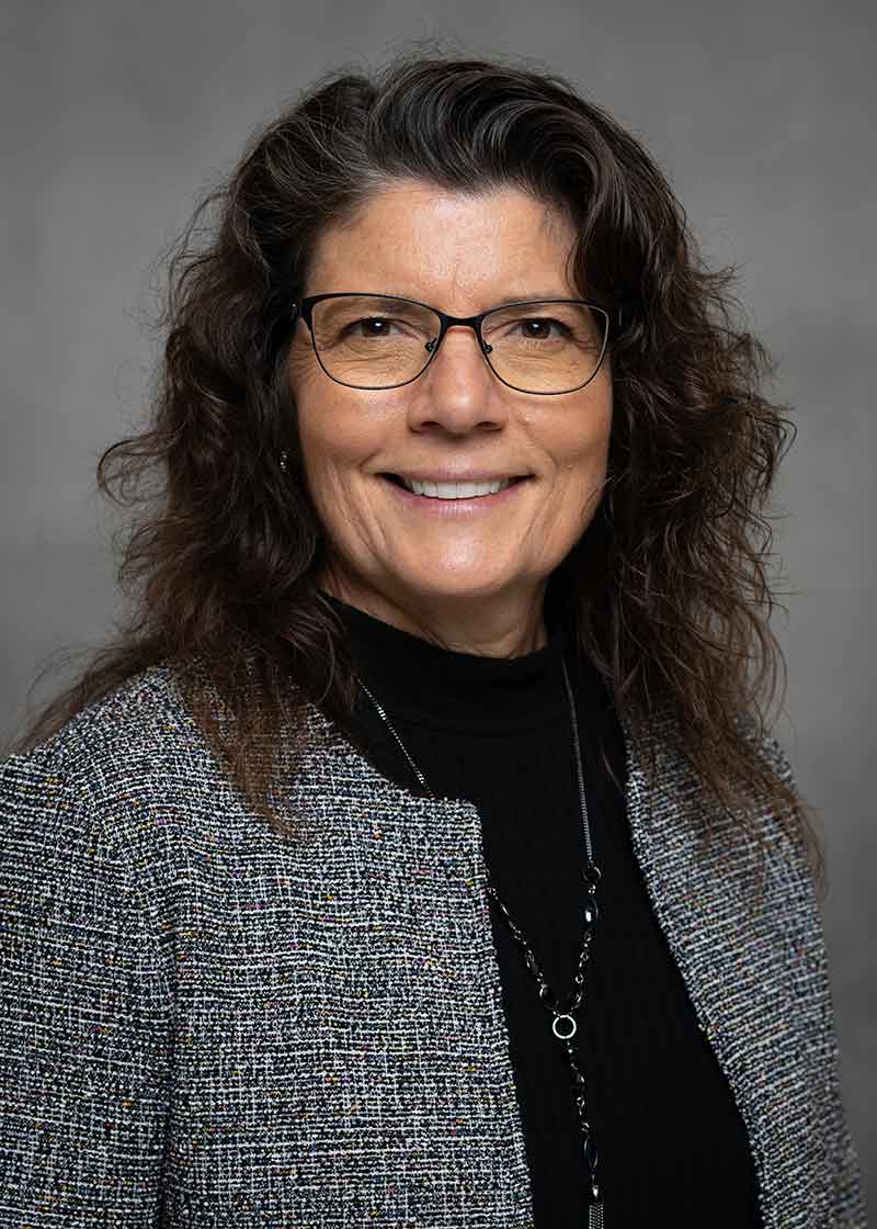 Headshot of a smiling woman in glasses, a black shirt and a grey blazer