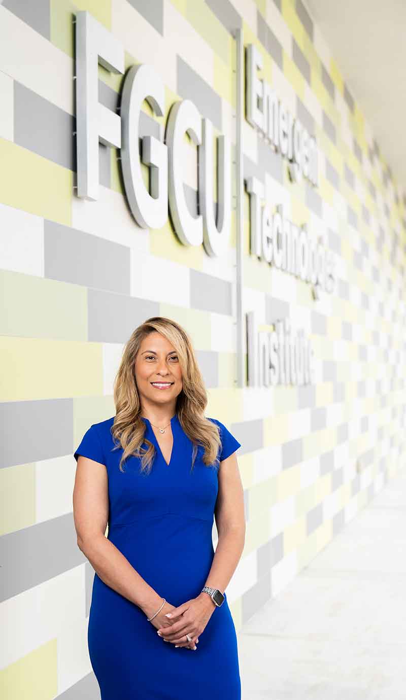 A smiling woman in a blue dress stands outdoors in front of a wall that reads FGCU Emergent Technologies Institute