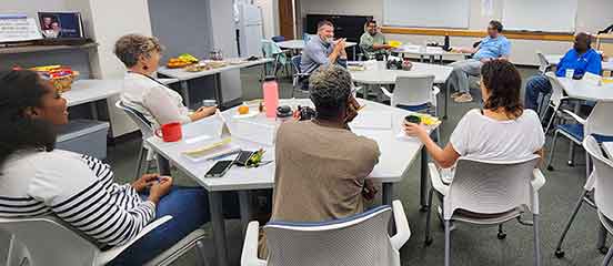 A group of people sit at tables with books in front of them, facing one man.