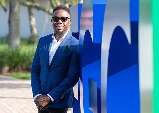 FGCU graduate Judes Sétalaire Albert standing in front of the FGCU logo on the Library Lawn on campus.