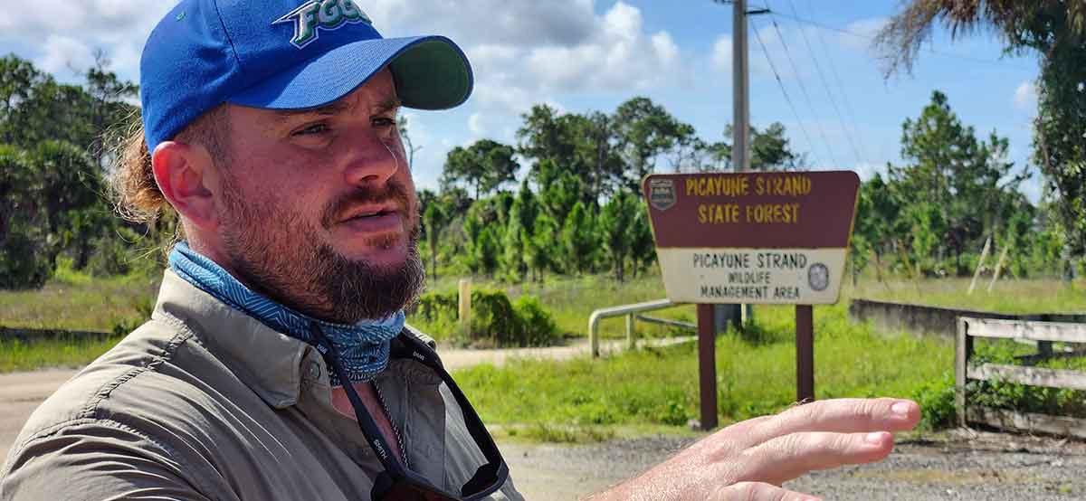 A man in profile wearing a blue FGCU hat and beige button-up shirt stands in front of a sign that reads Picayune Strand State Forest, Picayune Strand Wildlife Management Area