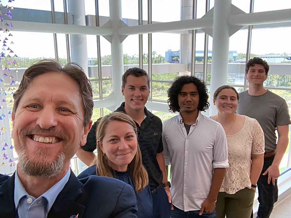 A group photo taken indoors of six people. The FGCU campus can be seen behind the group through a series of windows.