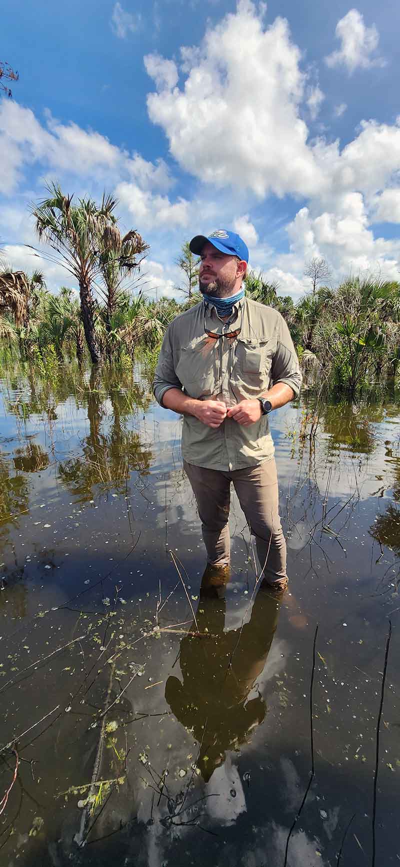 A man in a blue FGCU hat, khaki pants and a beige button-up shirt stands ankle-deep in water, with a few palm trees and saw palmetto visible behind him