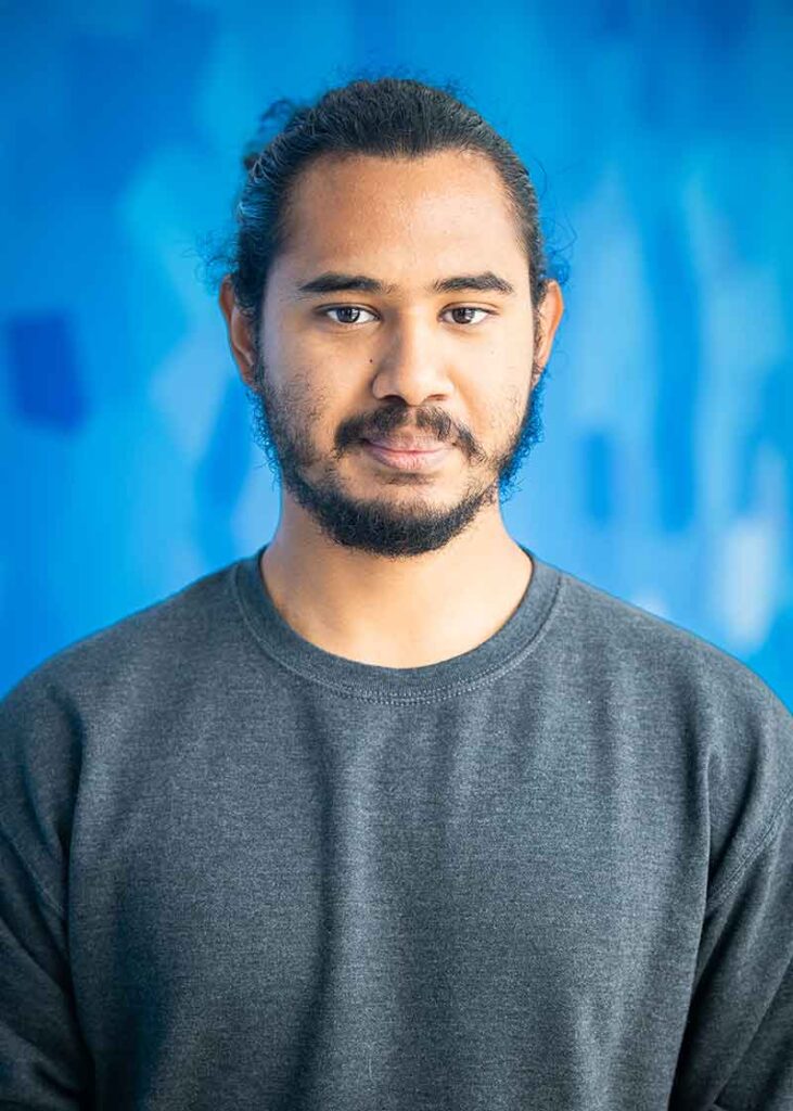 A headshot in front of a blue background of a young man in a grey crew neck top