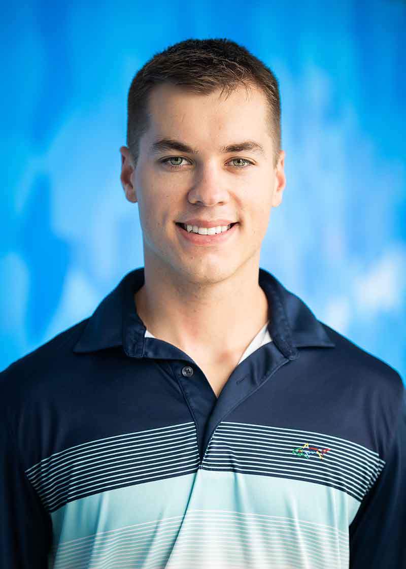 A headshot in front of a blue background of a young man in a navy, patterned and light blue polo