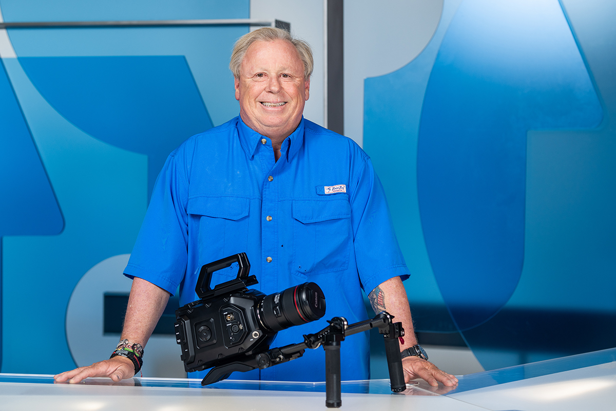 Man in blue shirt with video camera on set of news show.