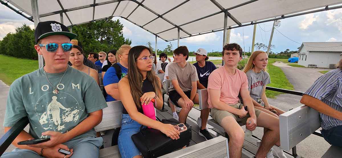 A group of roughly two dozen students sit in a covered trailer bed. There are trees and a blue sky with white clouds in the background.