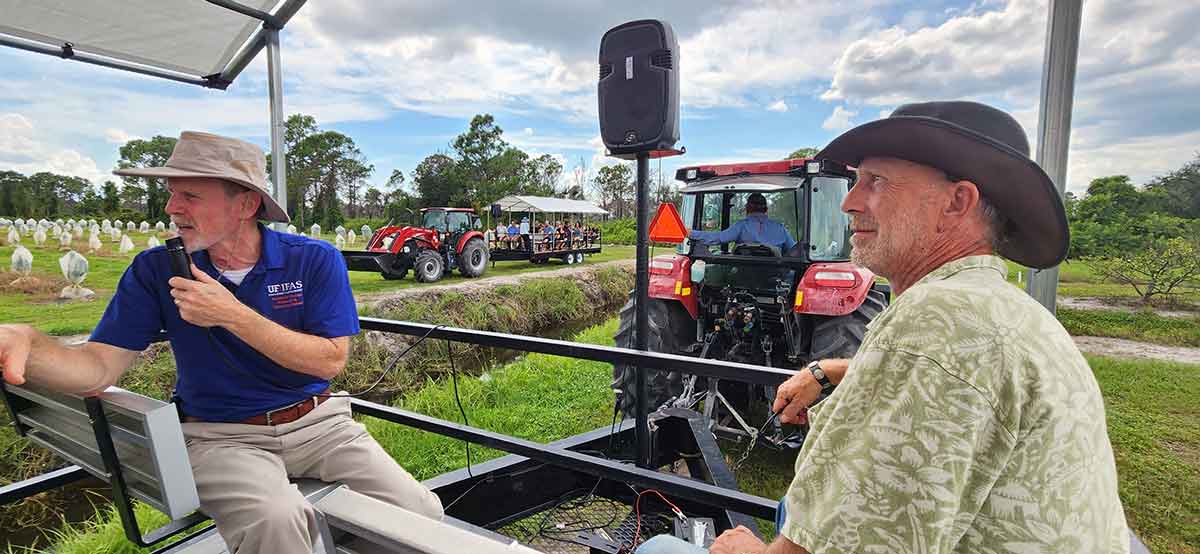 A man in a blue polo shirt holds a microphone and sits in a trailer being pulled behind a big, red tractor, next to a man in a cowboy hat and green shirt. In the distance, another tractor pulls another trailer through a field with two dozen passengers.