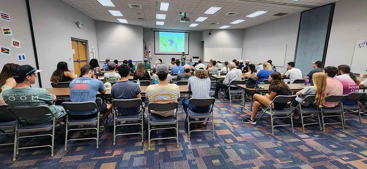 Inside a classroom, a woman uses a PowerPoint presentation as she stands near a podium. Roughly 50 people sit in metal folding chairs with their back to the camera.