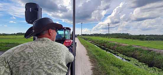 A man in a cowboy hat and green shirt, seated behind a big red tractor, looks our at a green farm as the clouds roll overhead.