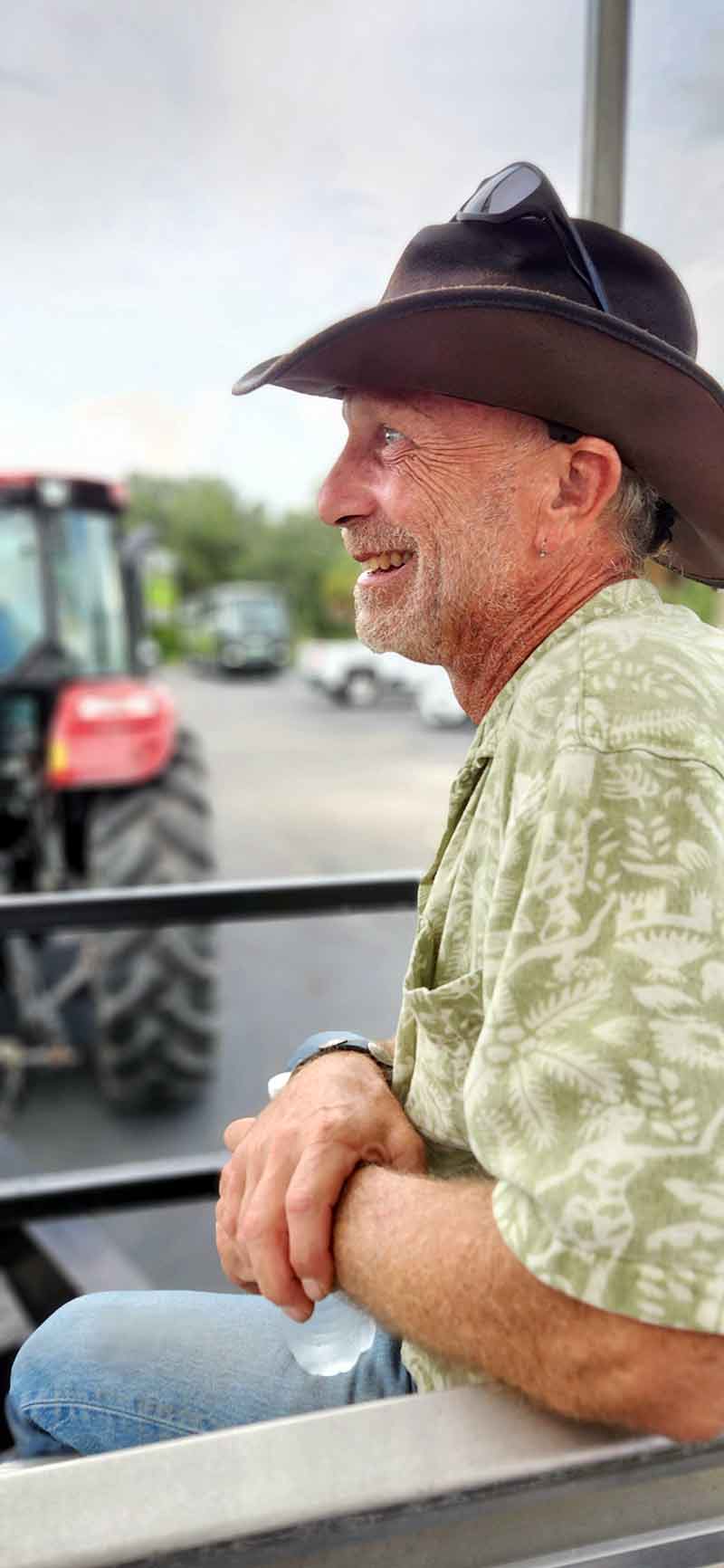 A man in a cowboy hat and green shirt sits smiling in a trailer behind a big, red tractor